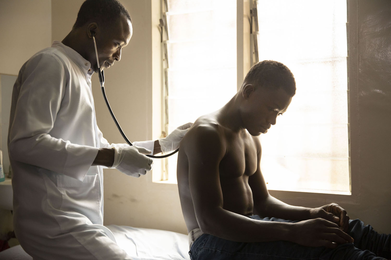 Healthcare Fotografie: In einem afrikanischen Krankenhaus untersucht ein Arzt einen jungen Patienten. Er hört mit dem Stethoskop die Lunge ab. Stimmungsvolle Gegenlichtaufnahme.