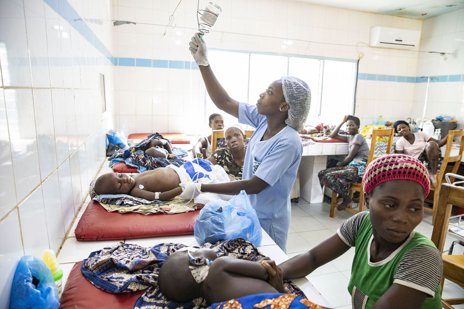 Healthcare Fotografie: in der Notaufnahme eines afrikanischen Krankrenhauses hängt eine Krankenschwester eine Infusion für einen kleinen Patienten an. Weitere Mütter mit ihren kranken Kindern sind im gleichen Raum zu sehen.